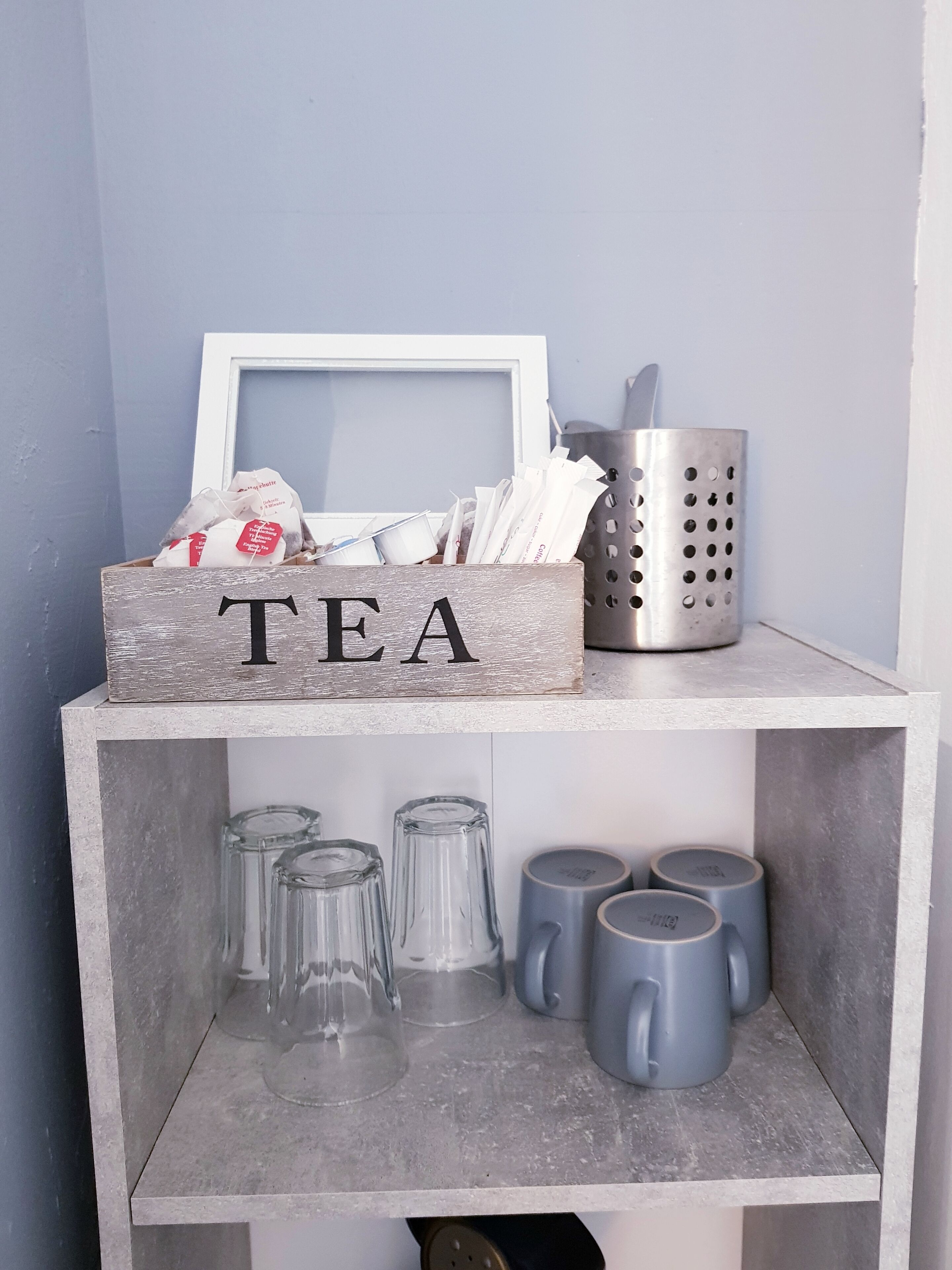 Une étagère sur laquelle se trouve une boîte en bois étiquetée « THÉ », contenant des sachets de thé, ainsi qu’un récipient en métal. En dessous, on peut voir plusieurs tasses en verre transparent et des tasses en céramique grise.