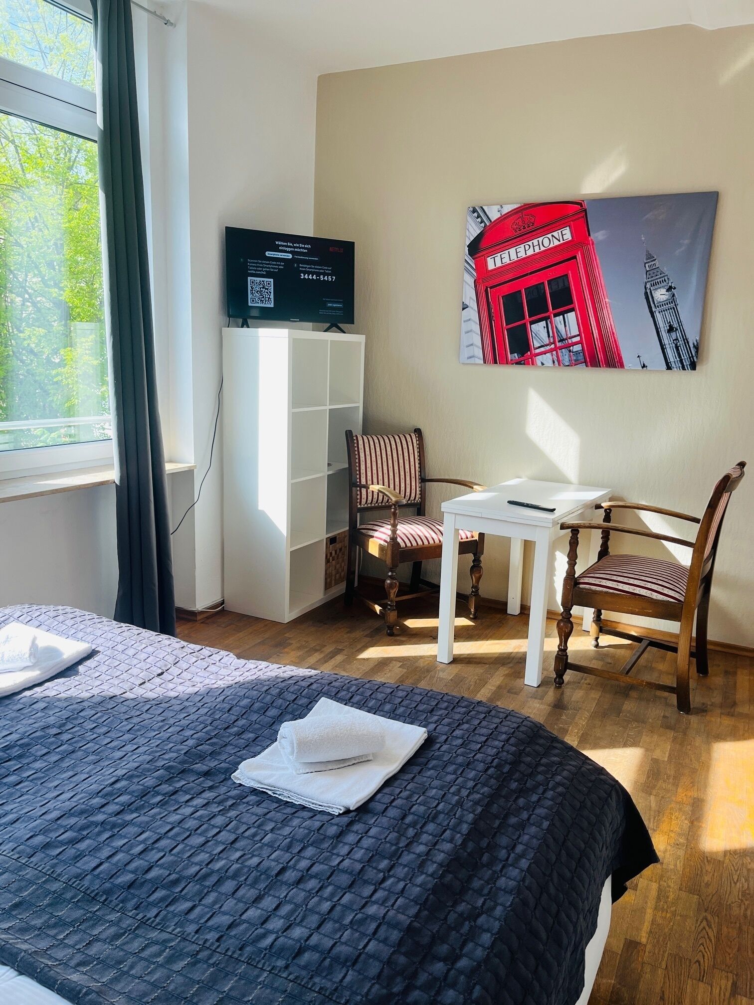 A hotel room with a bed, a desk, a chair, a television, and a painting of a red building.