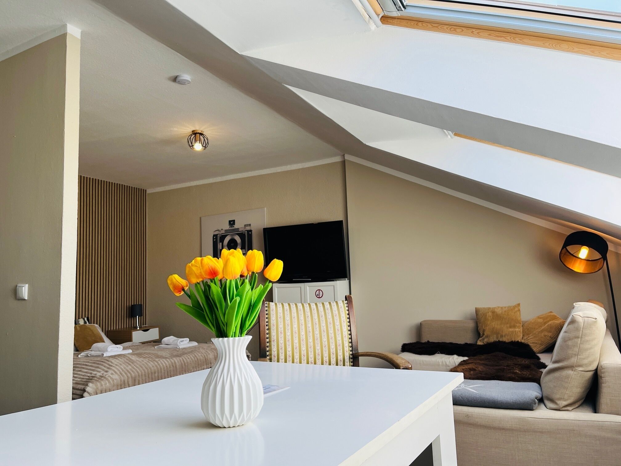 A modern living room with a white table, a vase of yellow tulips, a sofa, and a television.