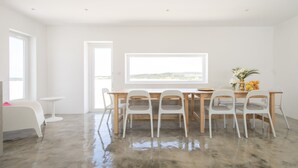 A modern, minimalist dining area with a wooden table and white chairs, large windows, and a view of the outdoors. - Casasupertubos (Peniche)