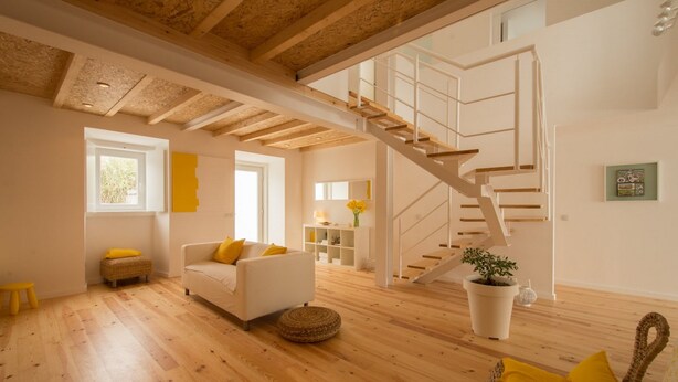 A spacious living area with a wooden floor, a white sofa with yellow cushions, a wicker ottoman, a potted plant, and a staircase leading to an upper level. - Casasupertubos (Peniche)