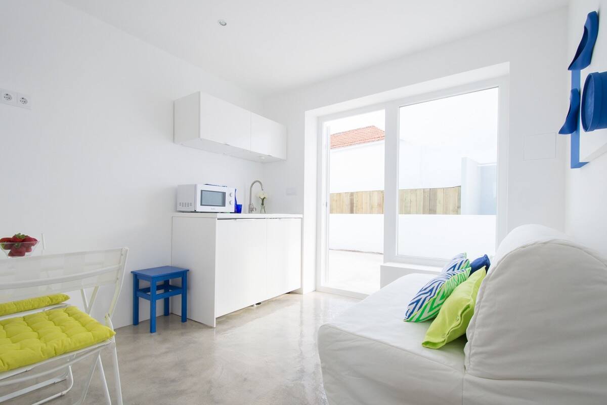 A modern, bright room with a white bed, a small kitchenette, and a large window.