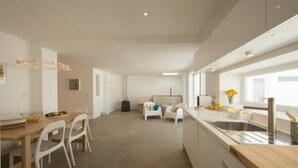 A modern kitchen with a large island, a dining area with white chairs, and a view of the outdoors through large windows. - Casasupertubos (Peniche)