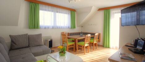 A modern living room with a grey sofa, green curtains, a dining table with wooden chairs, and a flat-screen TV.