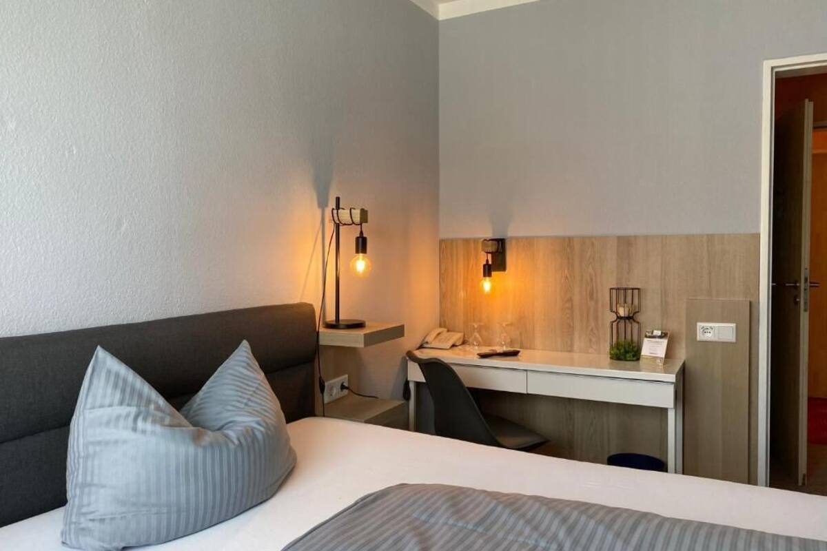 A hotel room with a bed, a desk, and a chair. There is a lamp on the desk and a phone on the wall.