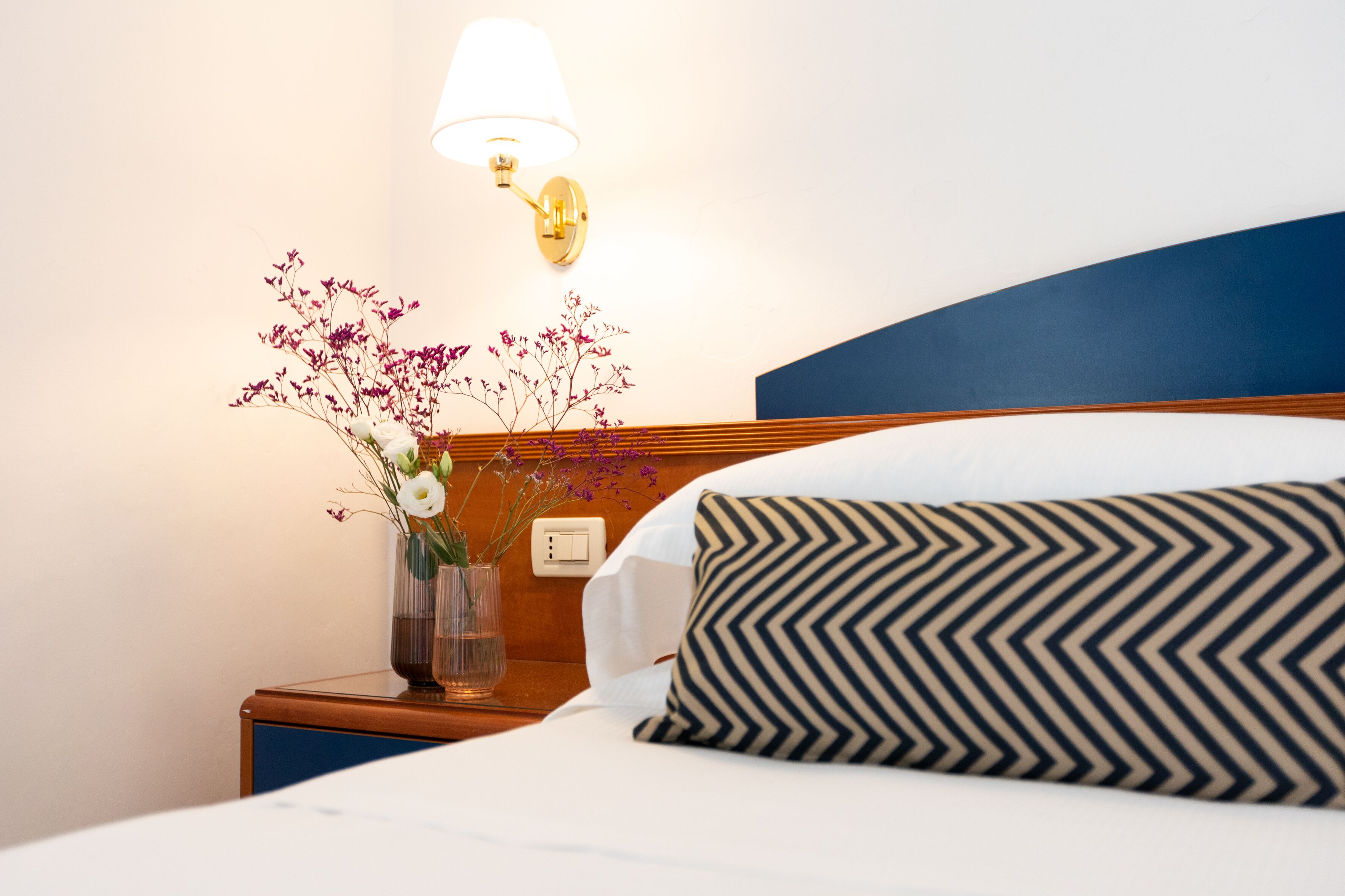 A neatly made bed with a patterned pillow, a bedside table with a vase of flowers, and a wall-mounted lamp.