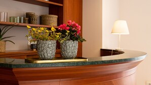 Un comptoir d’accueil au design incurvé en bois, deux plantes en pot ornées de fleurs jaunes et roses, et une lampe sur la droite.