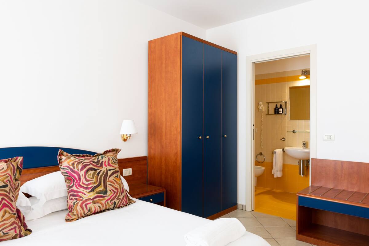 Habitación de hotel con cama, armario, mesita de noche y baño con lavamanos y espejo.