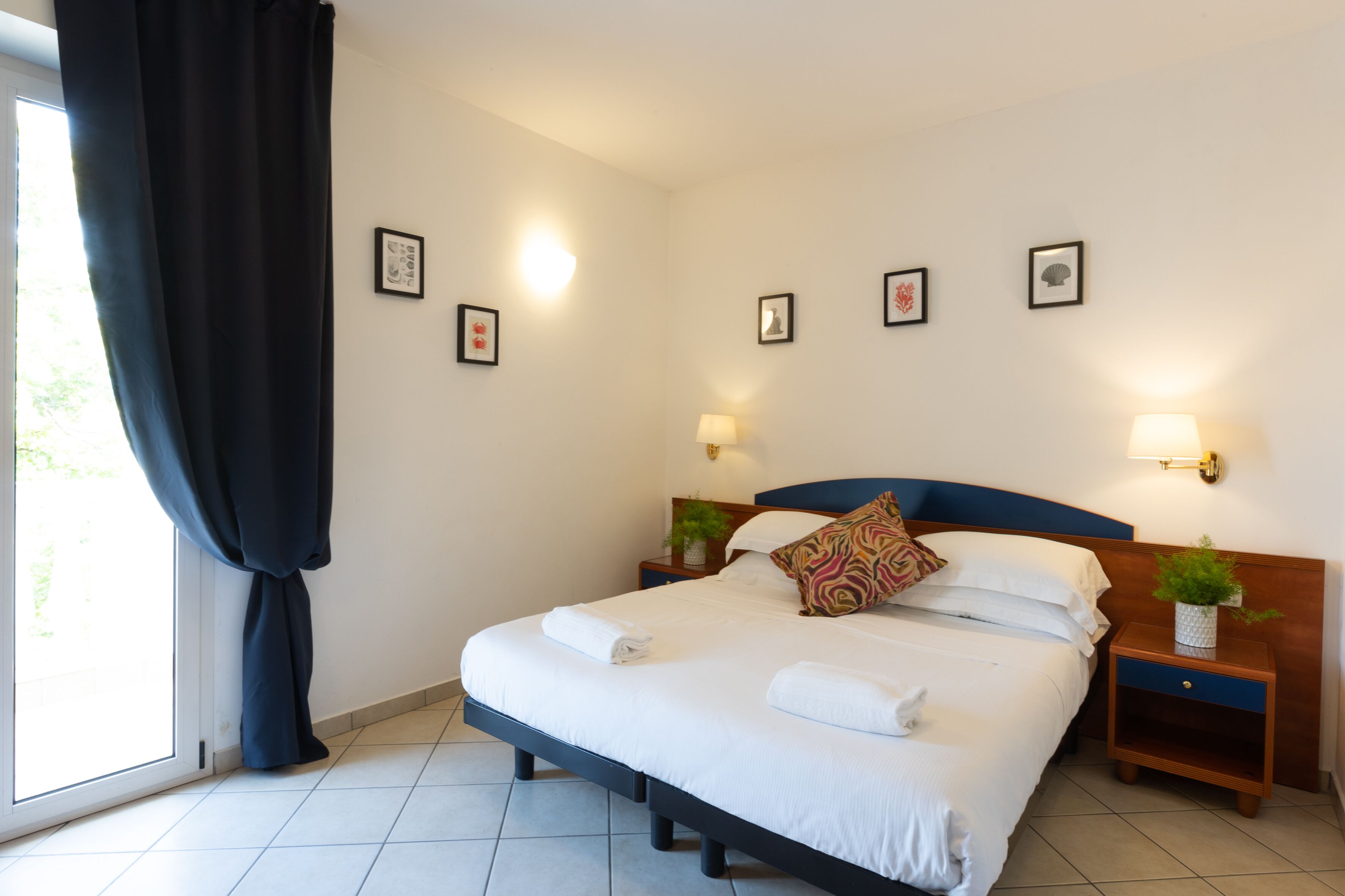 A neatly made bed with white linens, a blue headboard, and a decorative pillow. There are two bedside tables with lamps and a small plant on one. The room has a large window with a dark curtain, and the walls are adorned with framed pictures.