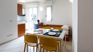 Un coin repas avec une table blanche, des chaises jaunes et un set de table bleu. On aperçoit en arrière-plan une cuisine avec des meubles en bois et un fourneau.