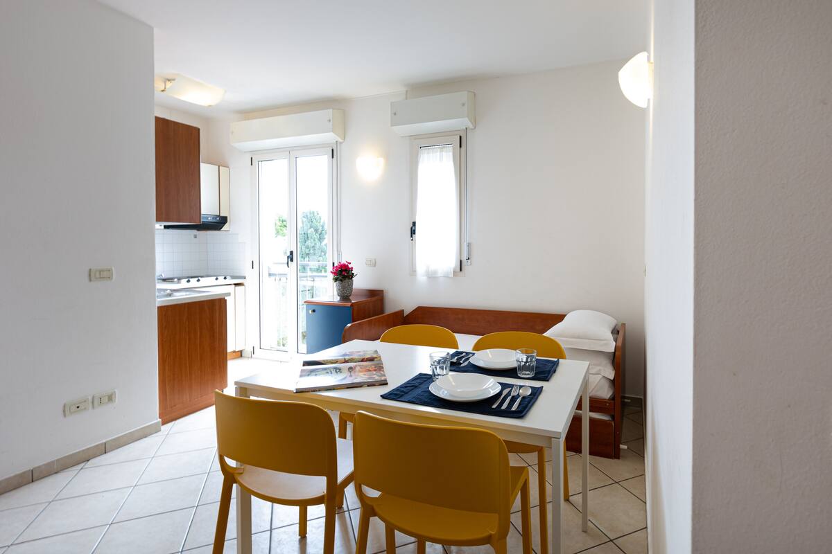 Un comedor con una mesa blanca, sillas amarillas y un mantel individual azul. Se observa una cocina al fondo con armarios de madera y una estufa.