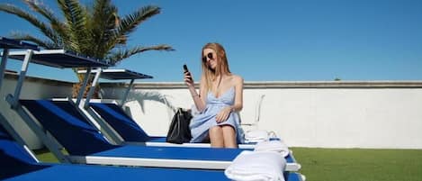 Una mujer sentada en una tumbona con un teléfono inteligente, un bolso negro y una toalla blanca en la tumbona de al lado.