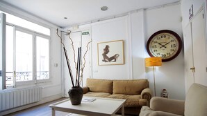 Ein Zimmer mit einem beigen Sofa, einem weißen Tisch mit einer Vase, einer Wanduhr und einer Tür mit einem Schild.