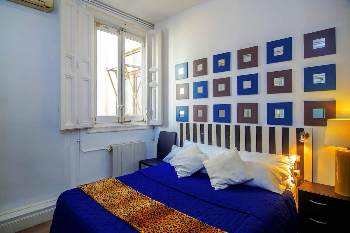 Une chambre à coucher avec un lit, une fenêtre, un radiateur, un mur orné de tableaux encadrés et une table de chevet avec une lampe.