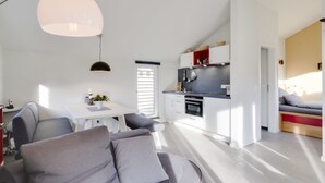 Cocina moderna con armarios blancos, encimera negra y estante rojo. Hay una zona de comedor con mesa blanca y sofá gris.