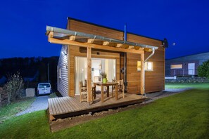 Une cabane en bois moderne, dotée d’une terrasse, avec une voiture garée à l’extérieur et un intérieur bien éclairé.