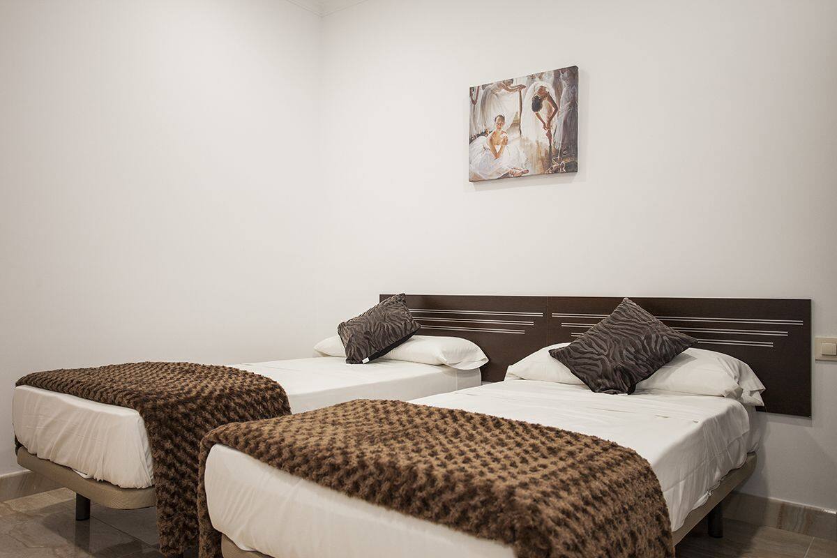 Hotellihuoneessa on kaksi sänkyä, joissa kummassakin on ruskea, pehmeä heittopeitto ja valkoiset vuodevaatteet. Sänkyjen yläpuolella seinällä on taulu.