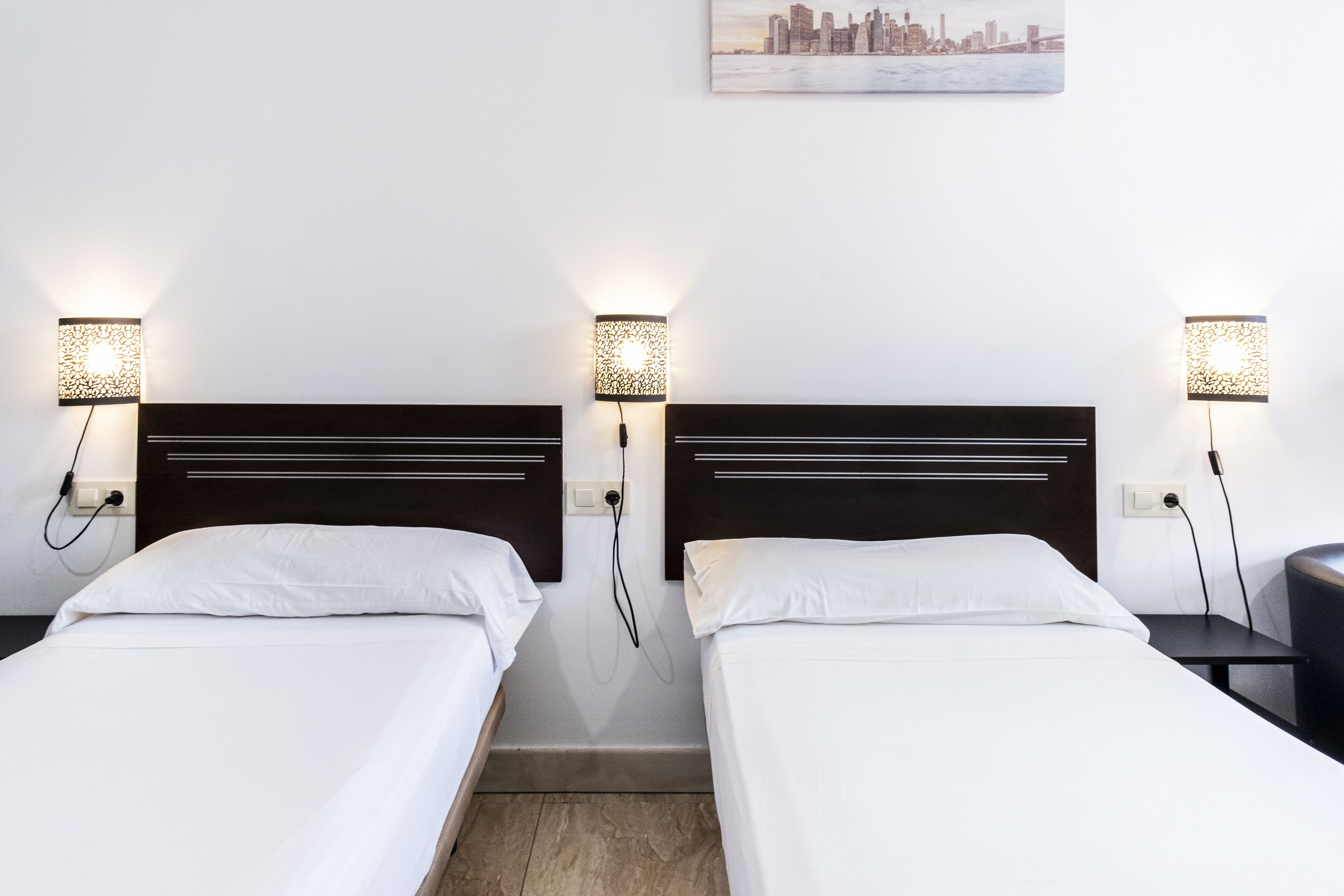 Two single beds with white linens, each with a wall-mounted lamp and a power outlet.