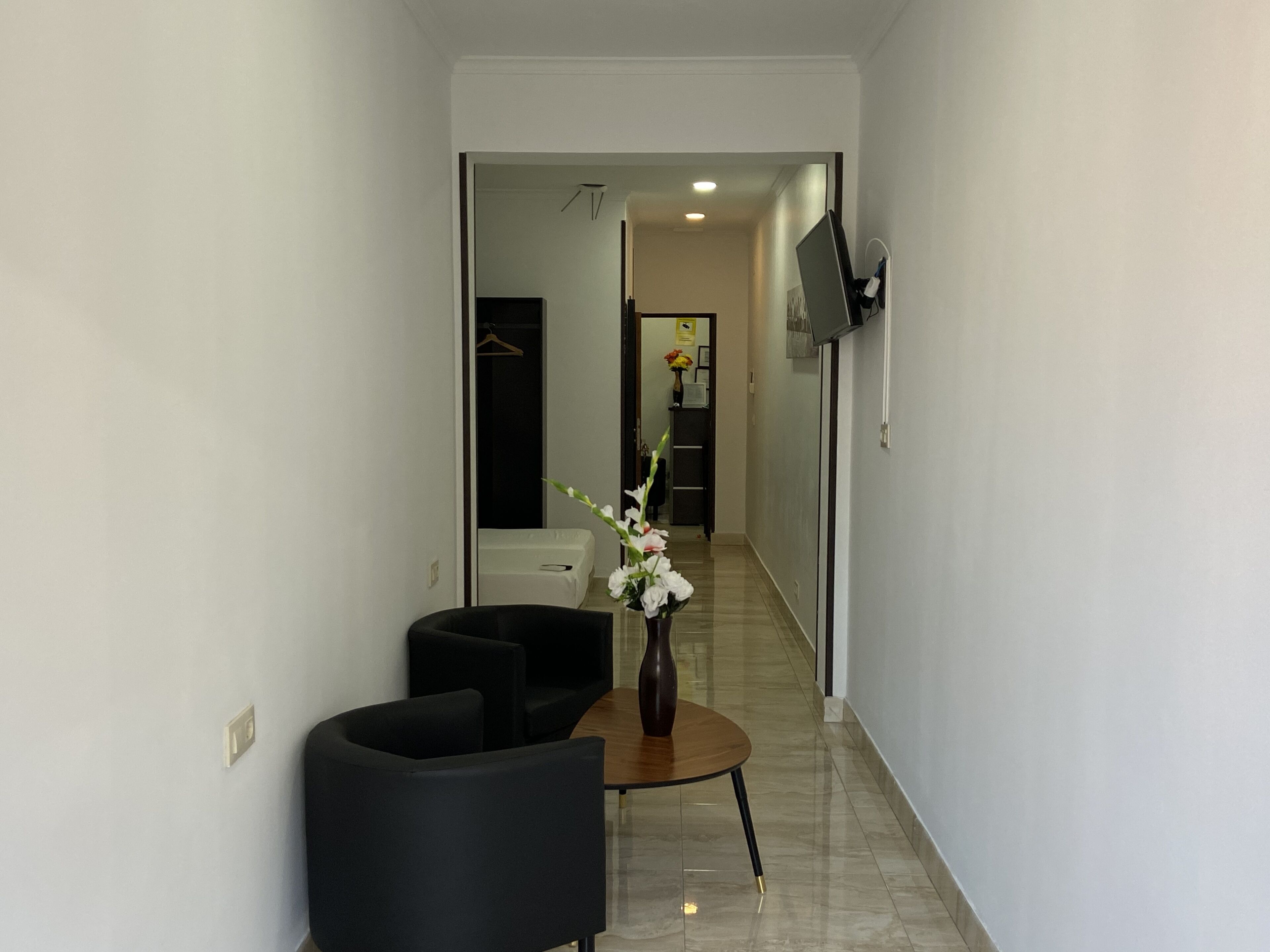 A hallway with a seating area featuring a round table and two chairs, a vase with flowers, and a television mounted on the wall.