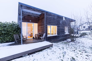 Une cabane moderne en bois, dotée d’une terrasse, de grandes fenêtres et d’un intérieur visible comprenant un coin repas.