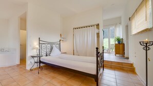A bedroom with a bed, a bedside table, a lamp, a window with curtains, a bathroom with a bathtub, and a staircase leading to an upper floor. - EIFELTRAUM Mediterrana - pure silence (Berlingen)