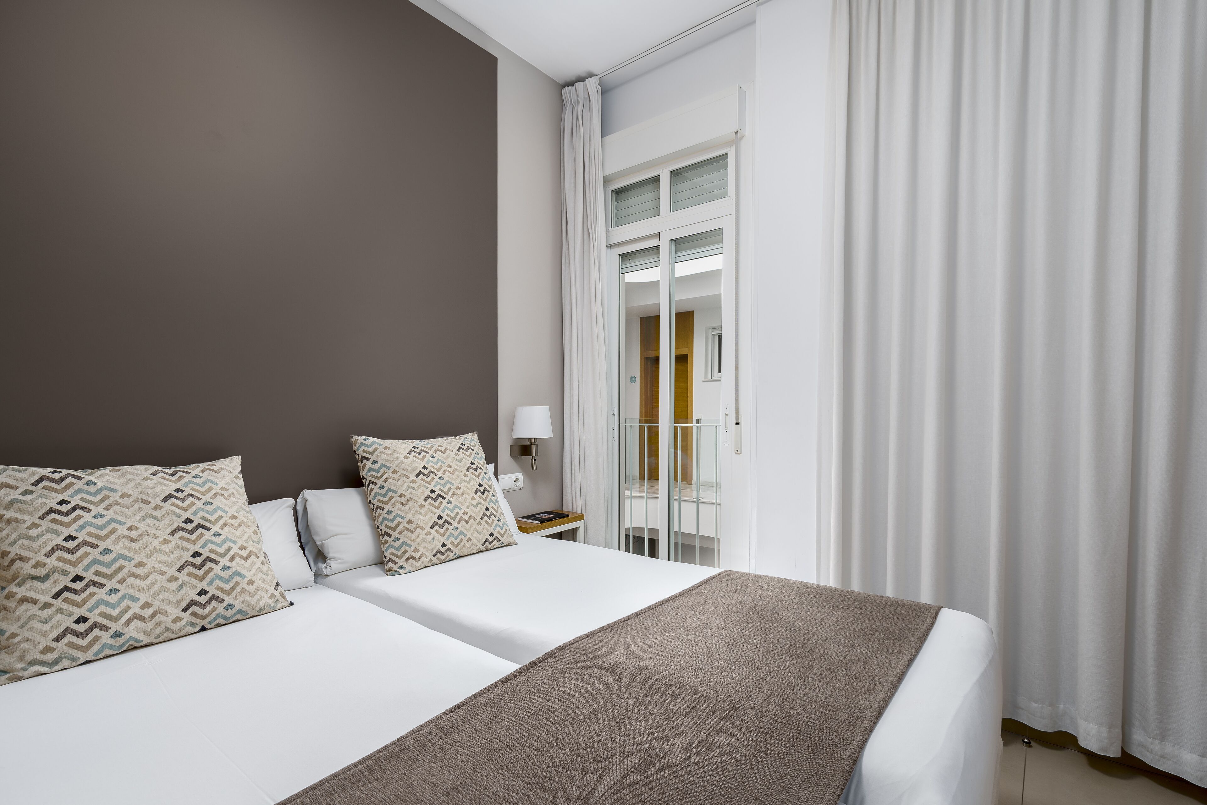 Habitación de hotel con dos camas, una mesita de noche, una lámpara y vista al exterior por una puerta.