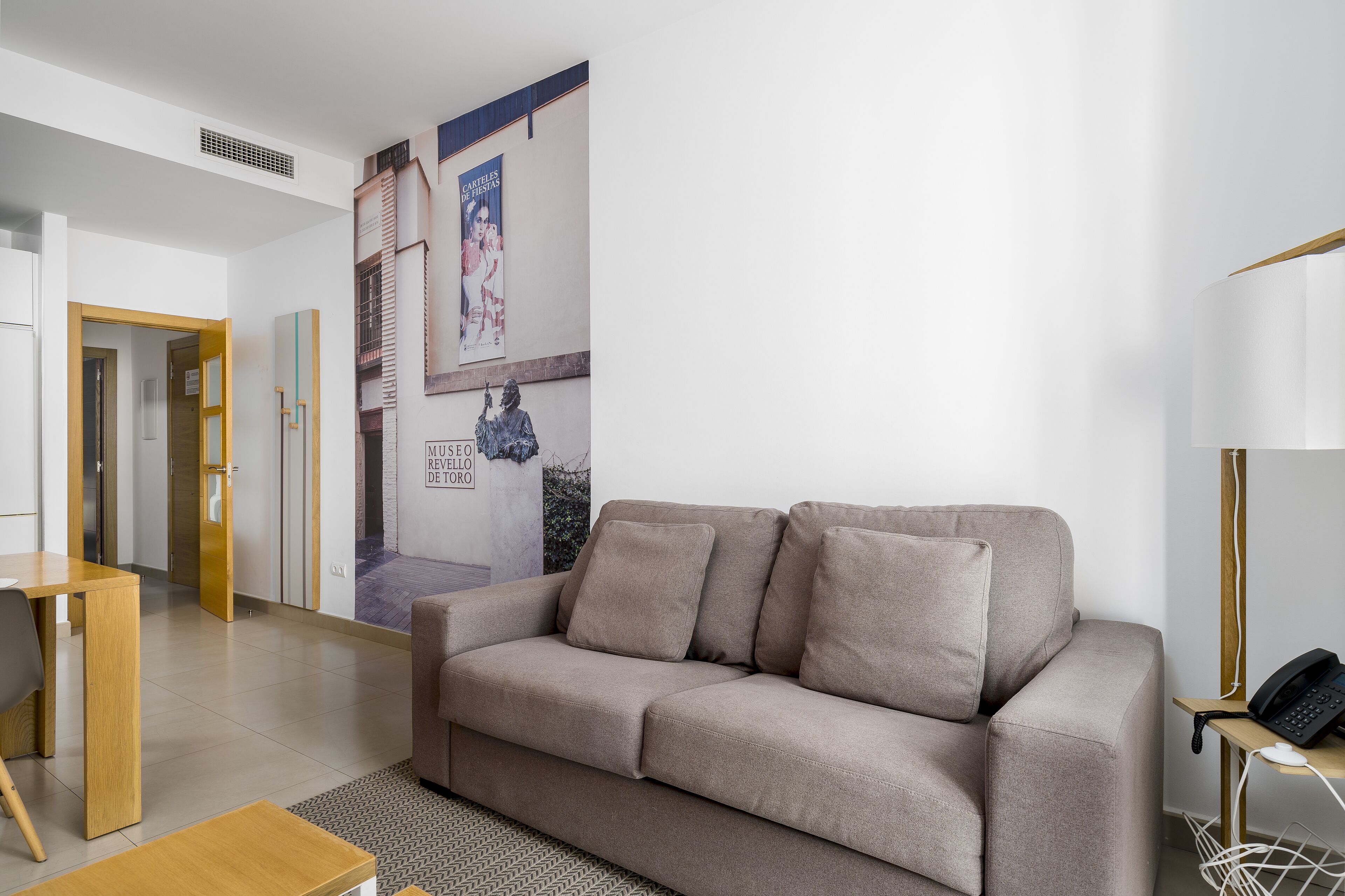 Ein modernes Wohnzimmer mit einem beigefarbenen Sofa, einem Holztisch und einem kleinen Beistelltisch mit einem Telefon. Ein Poster hängt an der Wand, und eine Tür führt in einen anderen Raum.