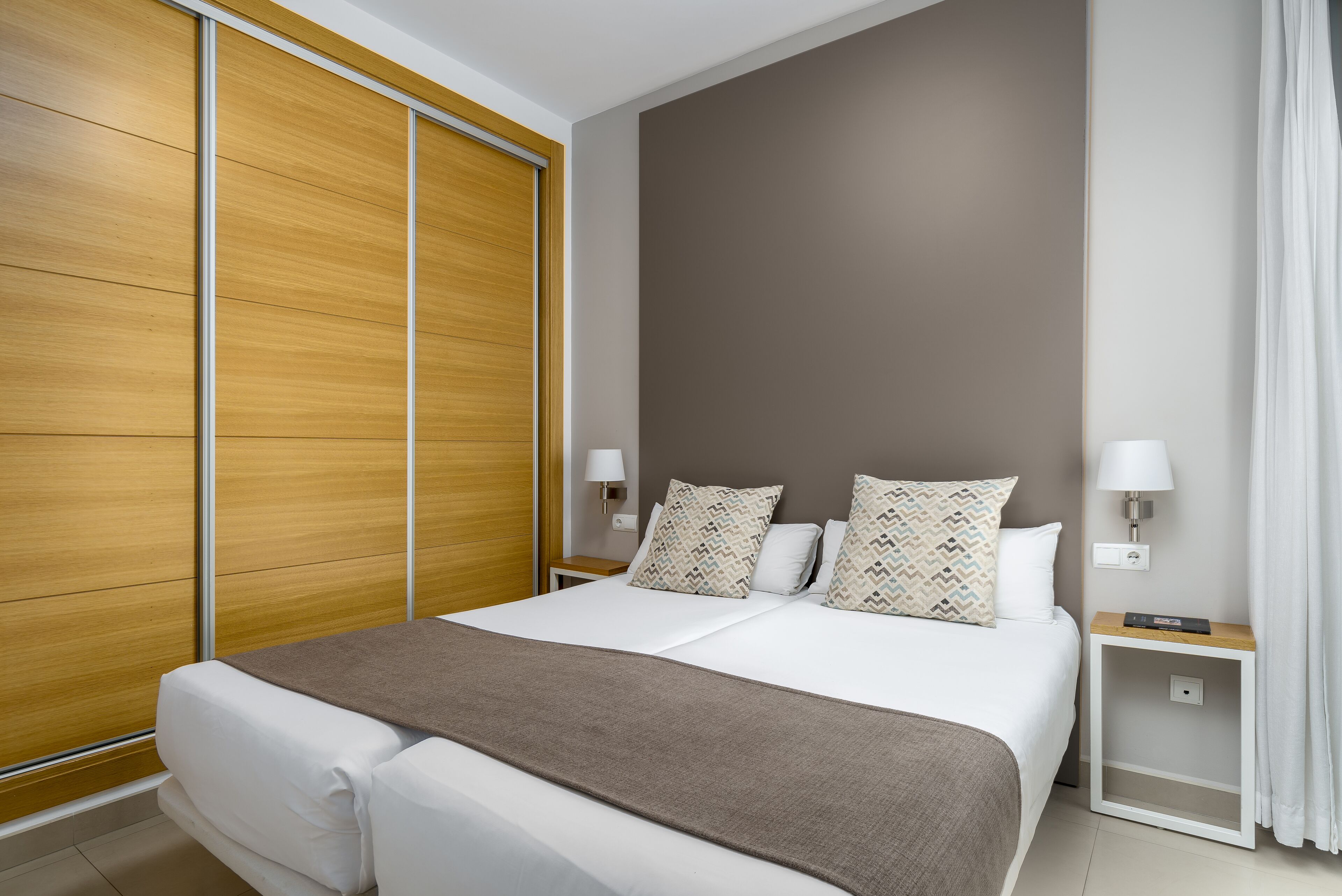 A modern hotel room with a large bed, wooden wardrobe, two bedside tables with lamps, and a neutral color scheme.
