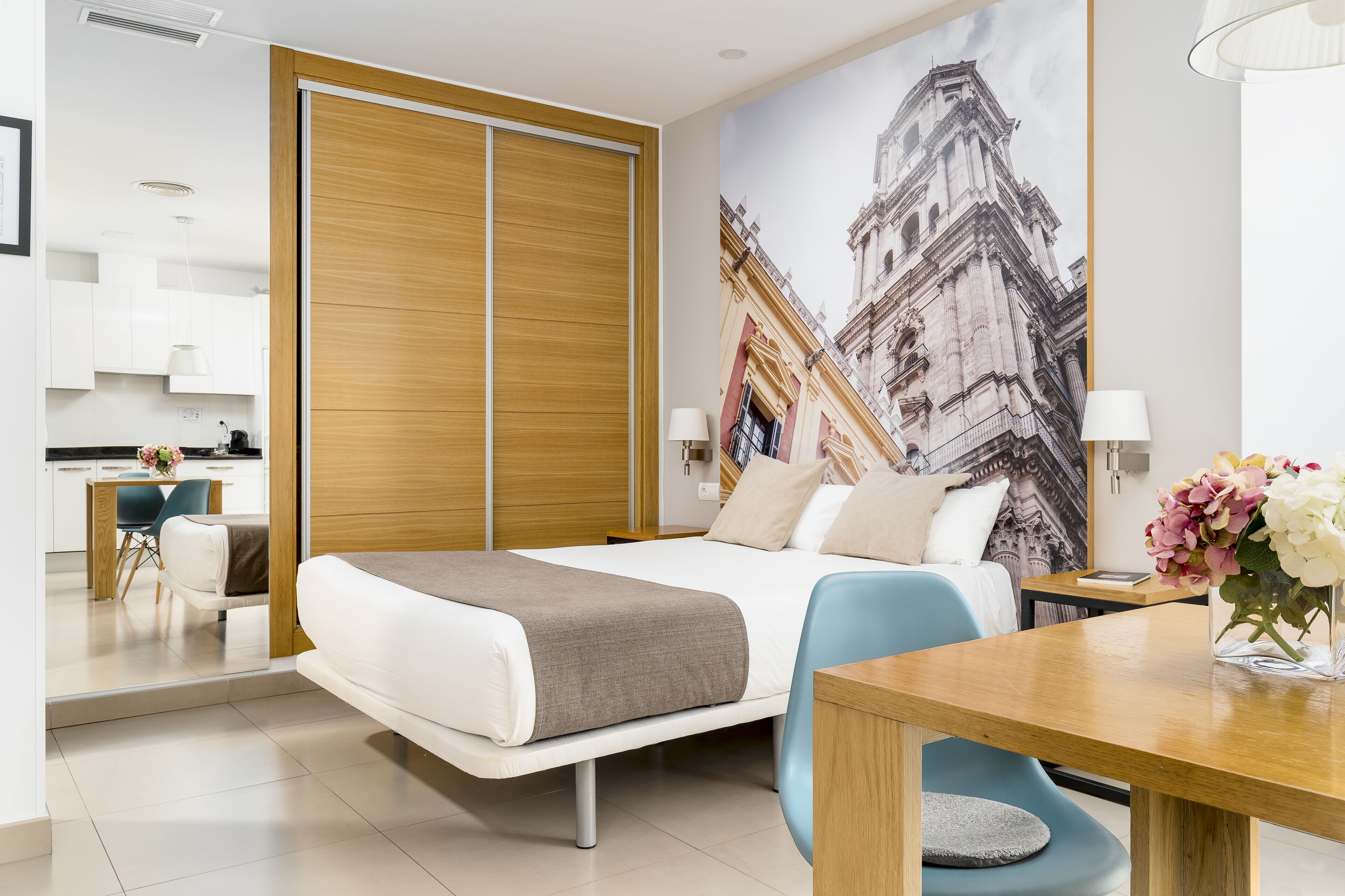 A modern hotel room with a bed, a dining table, a chair, a desk, a lamp, and a vase with flowers.