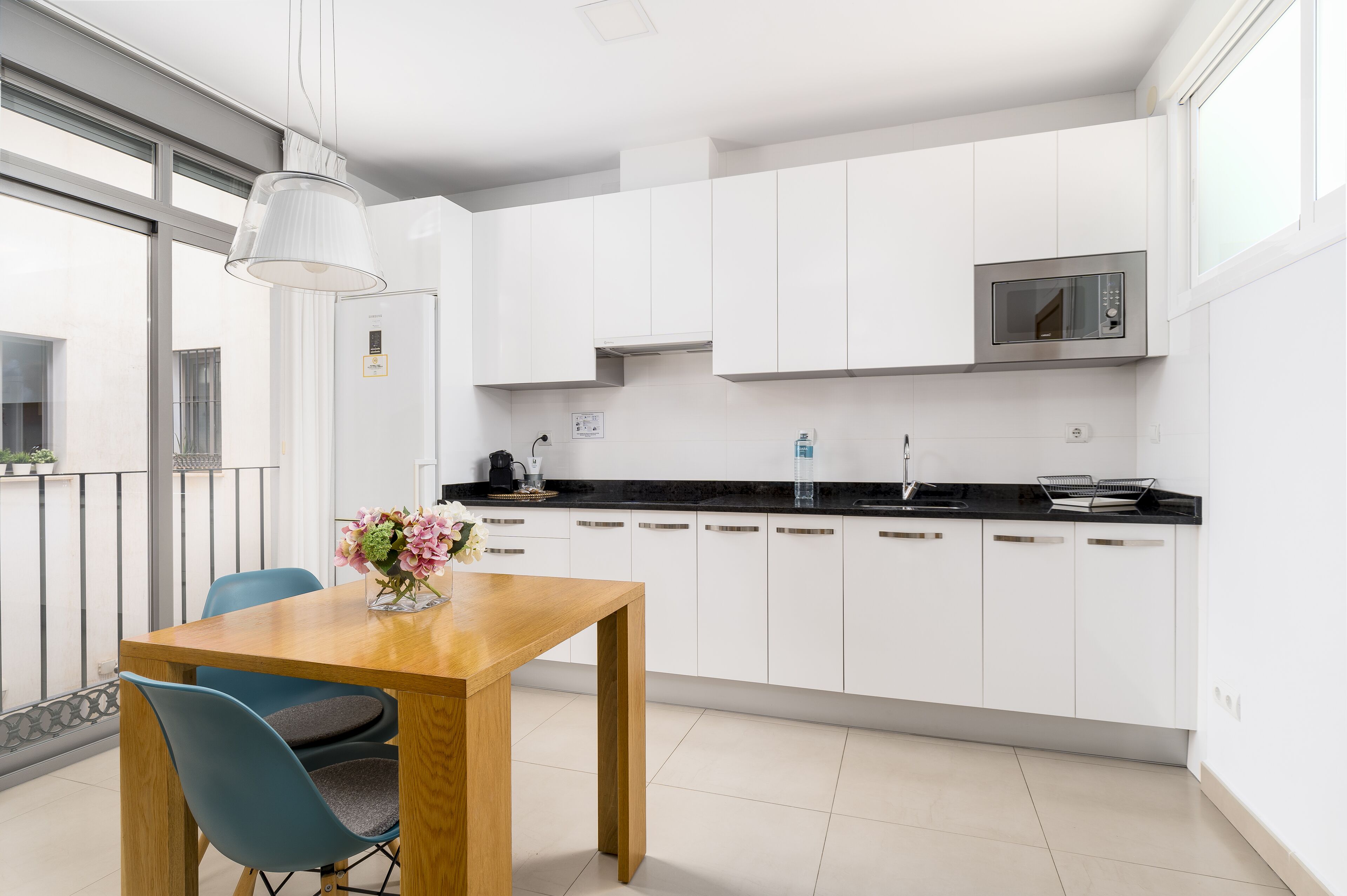 Una cucina moderna con mobili bianchi, un piano di lavoro nero e un forno a microonde in acciaio inossidabile. C'è un tavolo da pranzo in legno con sedie e un vaso di fiori.