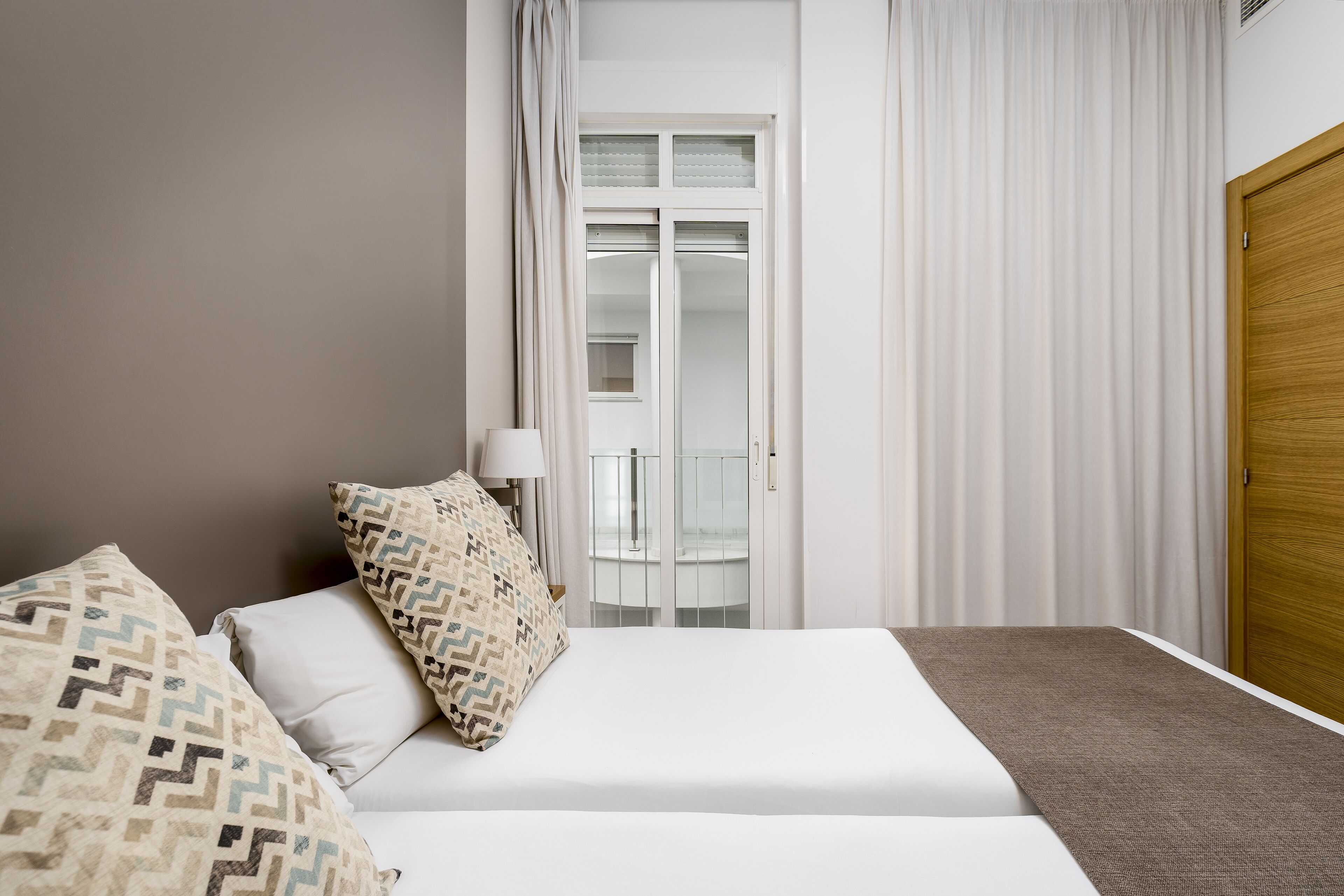 A bedroom with a bed, pillows, a nightstand, a lamp, a window with curtains, and a wooden door.
