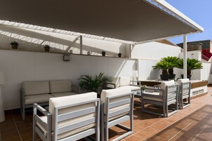 A covered outdoor seating area with a sofa and chairs, potted plants, and a tiled floor.