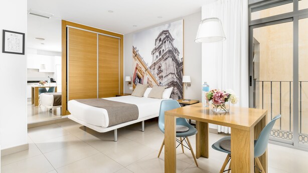 A modern hotel room with a bed, a dining table, and a view of a cityscape through a large window. - Suites del Pintor Historial Center, by Hoteles con Cultura (Málaga)