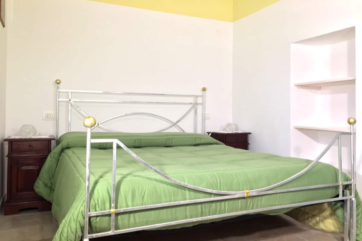 Una cama con estructura metálica y ropa de cama verde, una mesita de noche de madera y una estantería blanca.