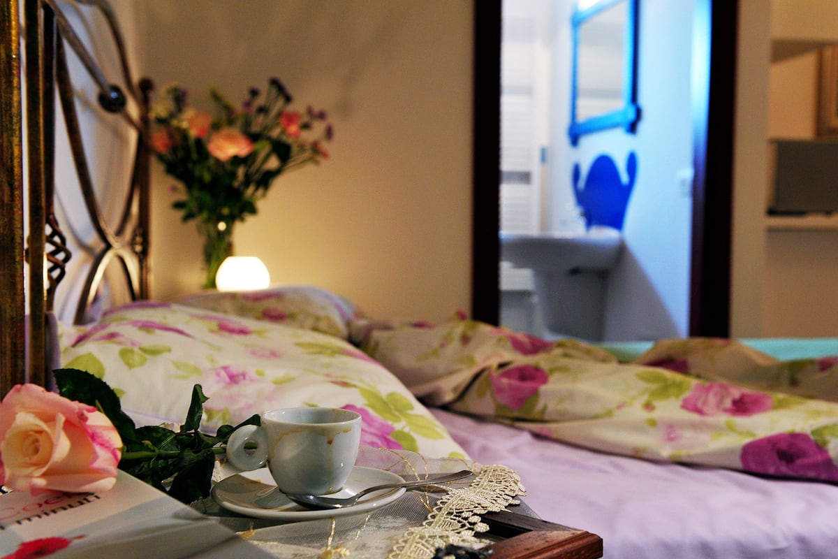 Una cama bien hecha con ropa de cama floral, una mesita de noche con una taza y platillo, un florero con flores, y un espejo que refleja un baño.
