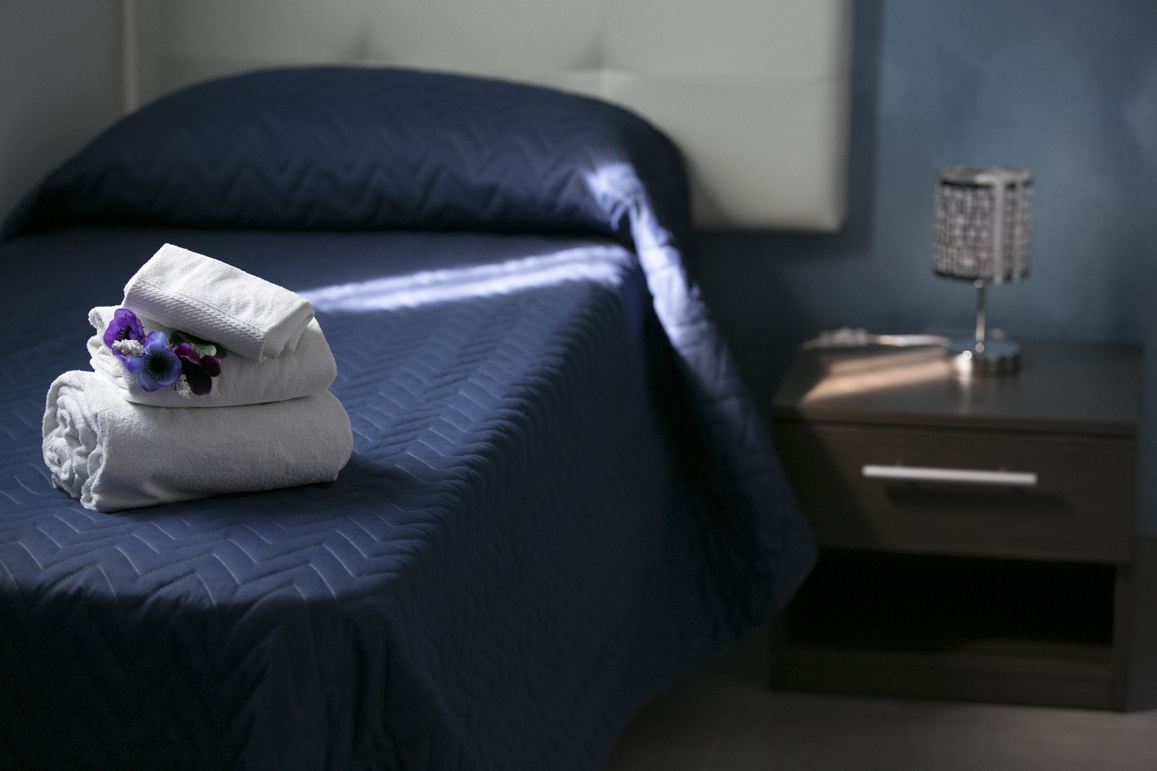 Een net opgemaakt bed met een opgevouwen witte handdoek en een paarse bloem, een nachtkastje met een lamp, en een donkerblauwe muur.