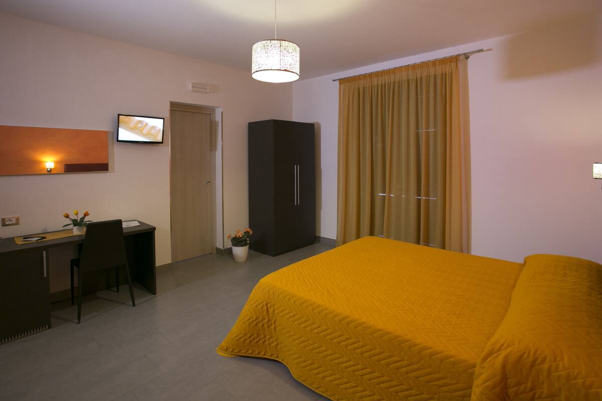 Habitación de hotel con cama, escritorio, silla, armario, planta en maceta y lámpara de pared.