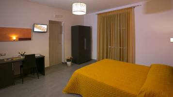 A hotel room with a bed, a desk, a chair, a wardrobe, a potted plant, and a wall-mounted light fixture.