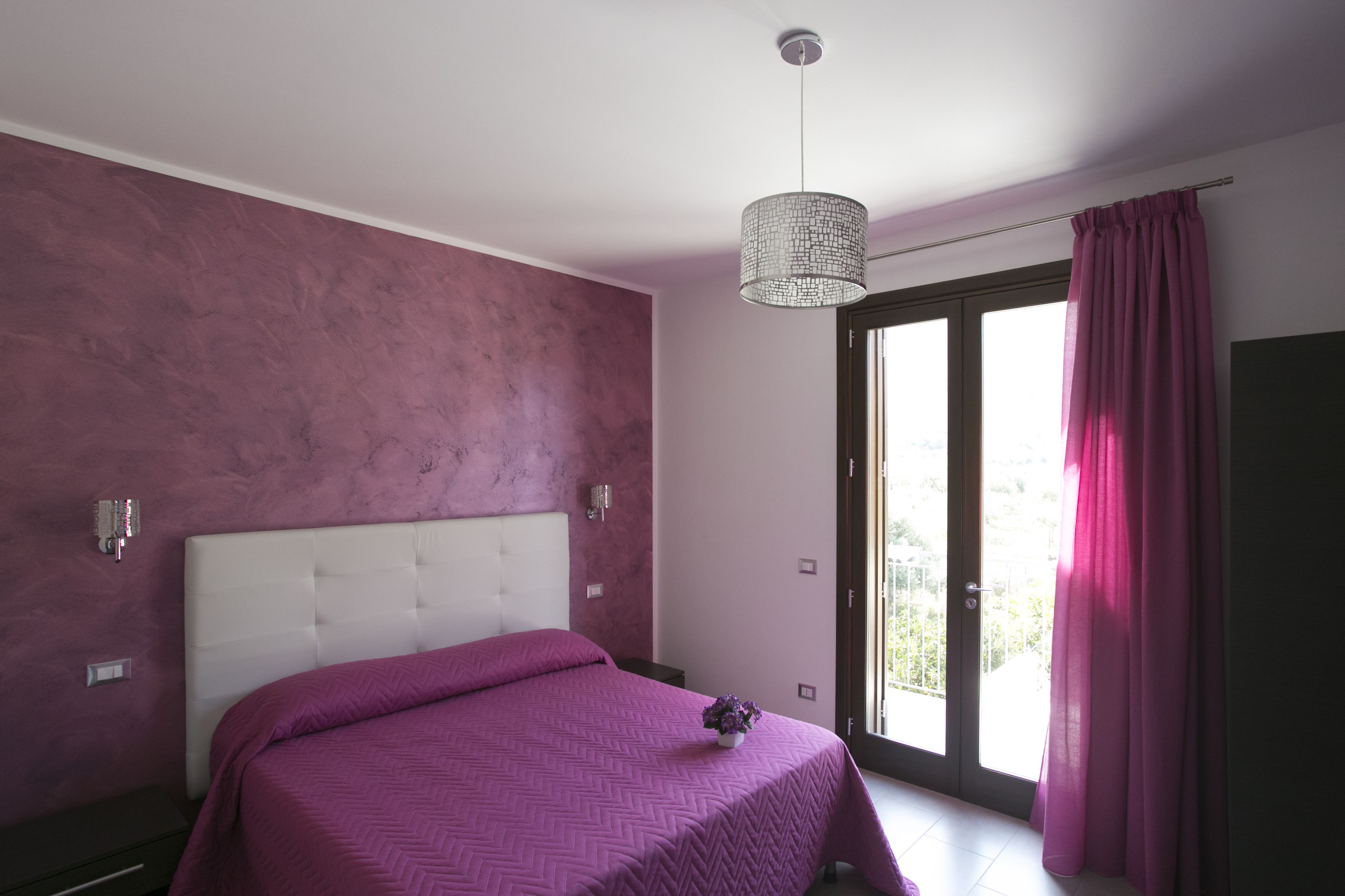 Ein Schlafzimmer mit einem großen Bett, einer Deckenleuchte und einer Glasschiebetür, die zu einem Außenbereich führt.