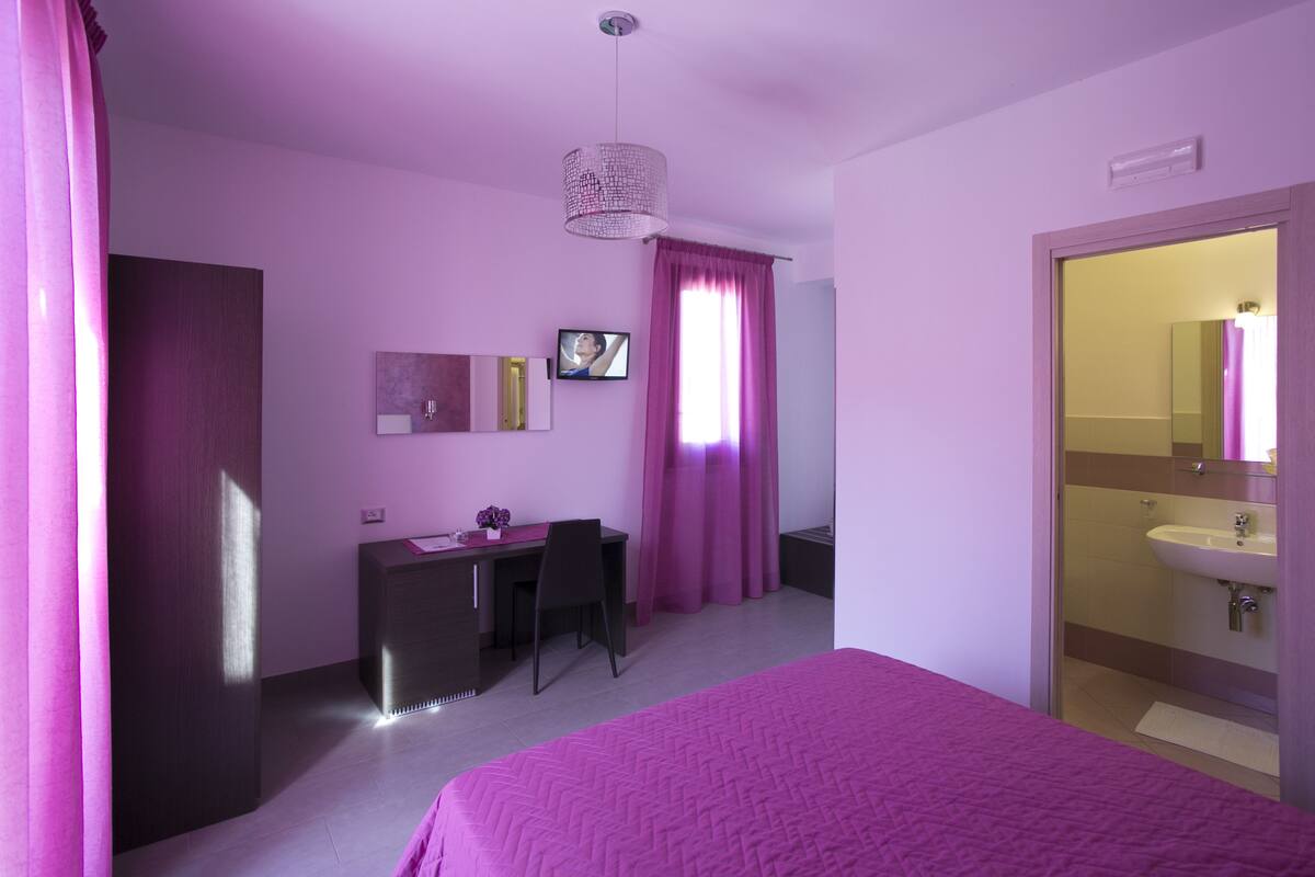 Un dormitorio con cama, escritorio, silla, televisor, espejo y baño con lavabo y ducha.