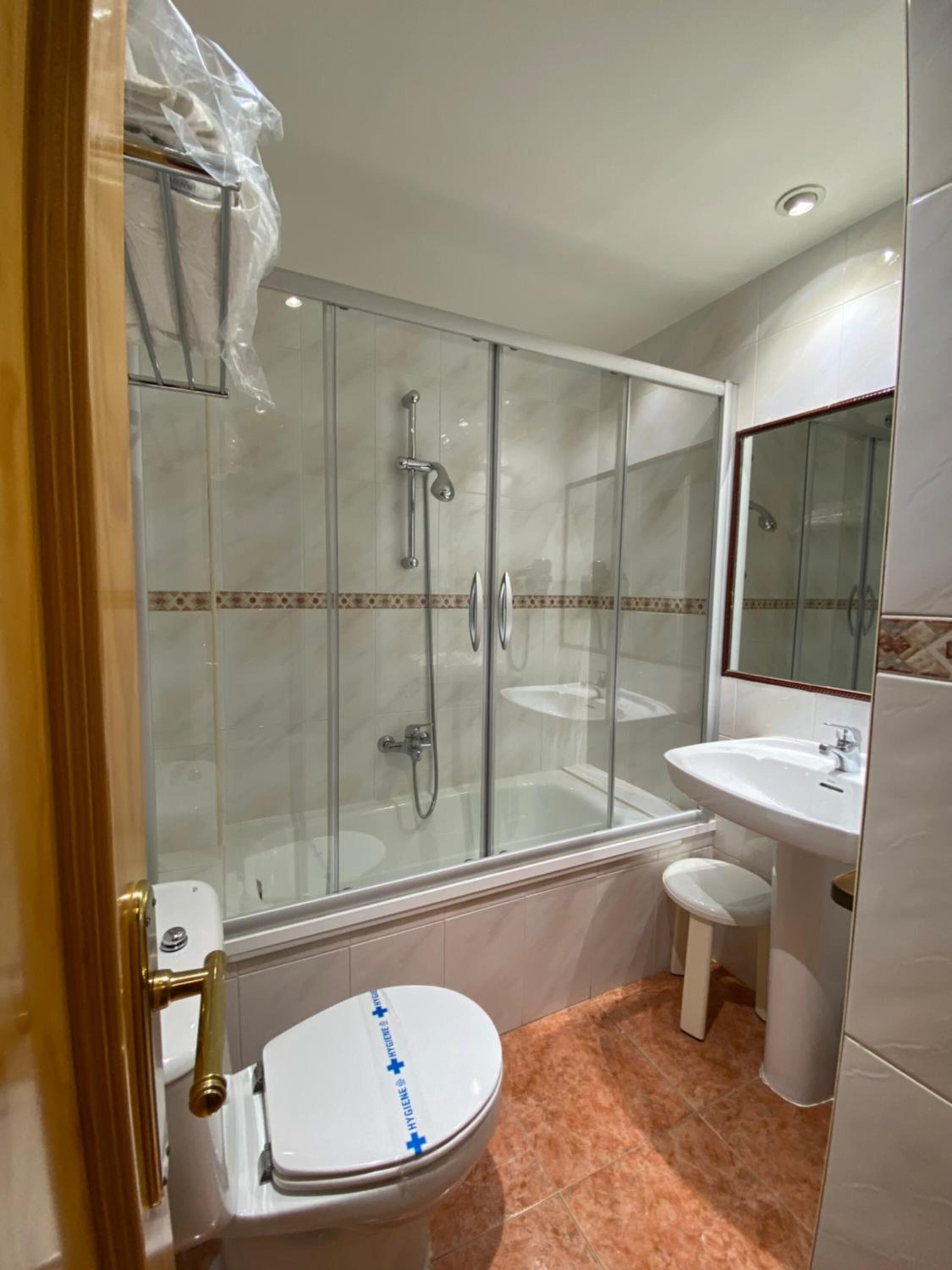 Un baño con ducha envuelta en vidrio, inodoro blanco y lavamanos pedestal.