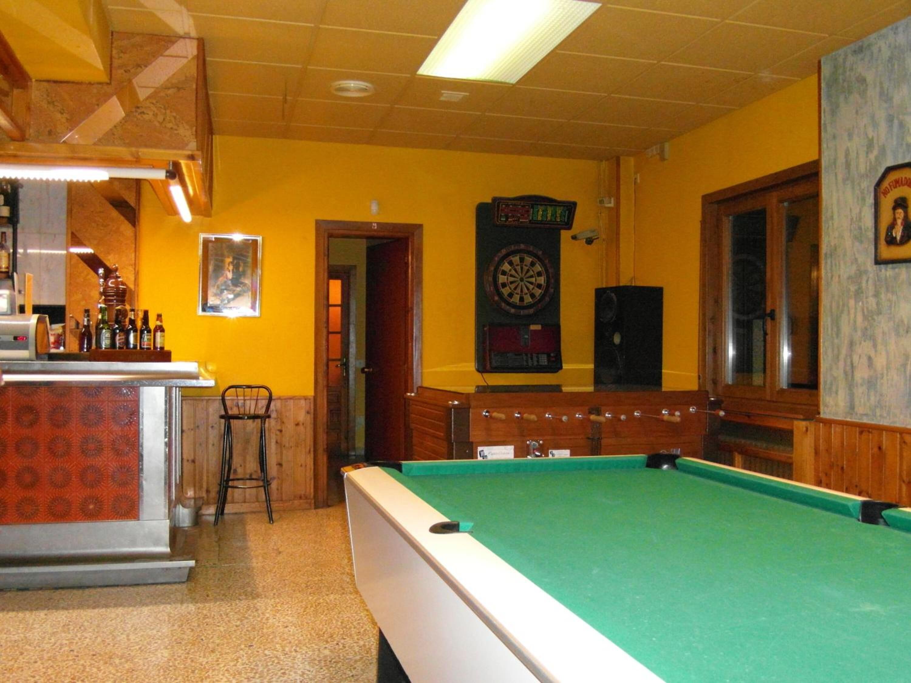 A pool table in a room with yellow walls, a dartboard, and a bar area with stools.