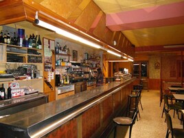 Una zona de bar con una barra larga, estanterías con botellas y vasos, y un espacio de estar con mesas y sillas.