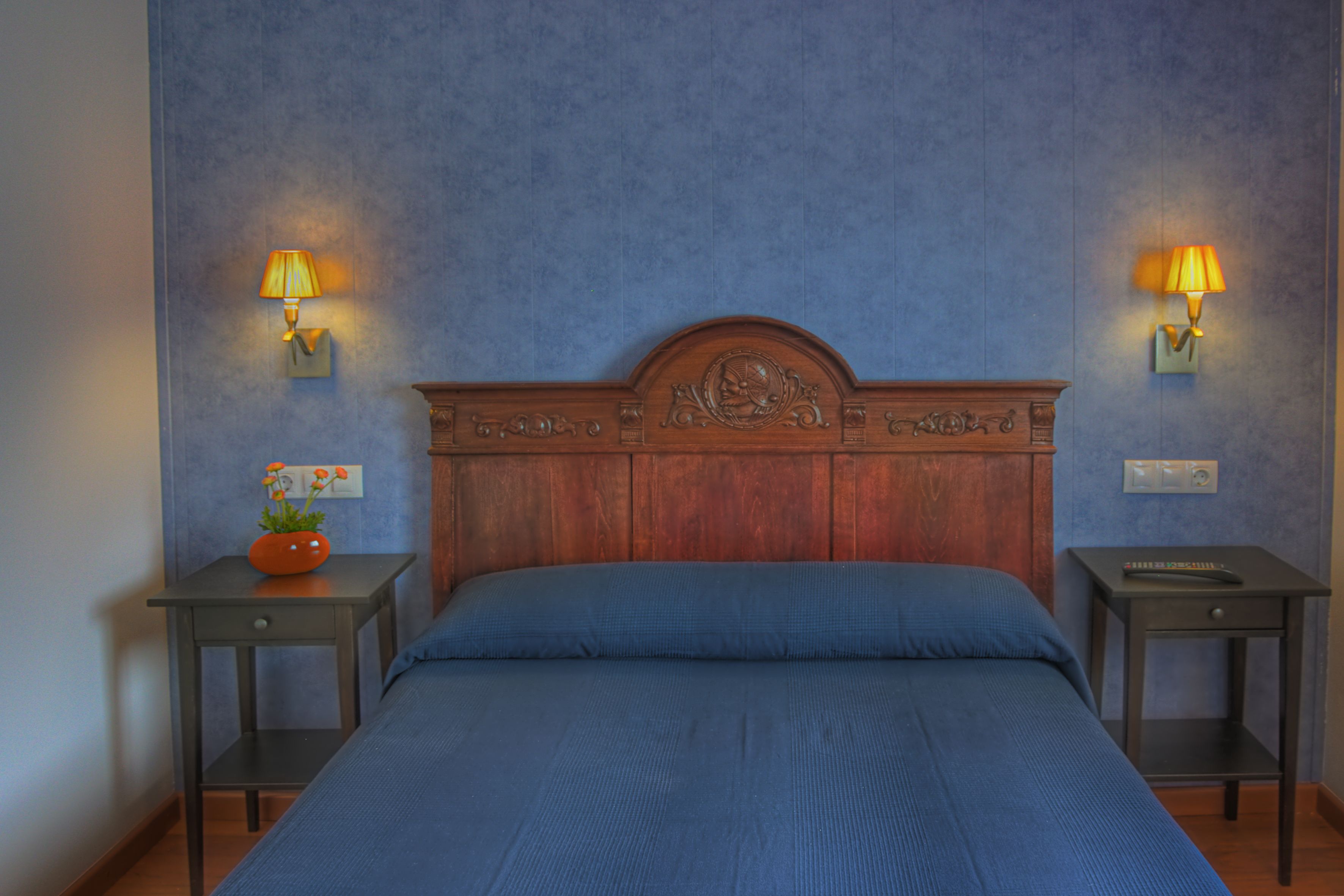 Un dormitorio con cabecera de madera, dos mesitas de noche, una colcha azul y dos lámparas de pared.
