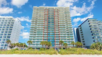 Un hotel moderno de gran altura con varios balcones, un pilar estructural rojo en el centro y vista a la playa.