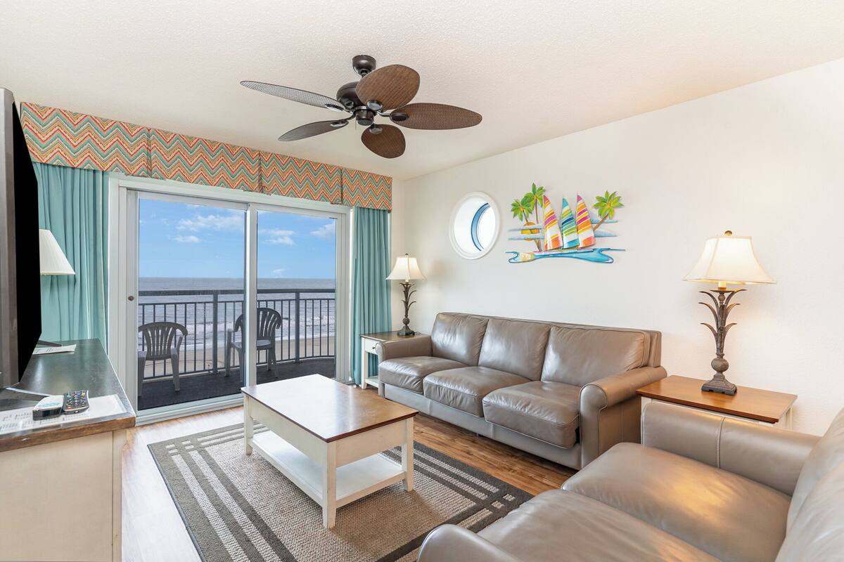 Ein Wohnzimmer mit einer Couch, einem Couchtisch und zwei Lampen. Es gibt einen Deckenventilator, ein Fenster mit Blick auf das Meer und eine Wand mit einer Uhr und einer Bootsdekoration.