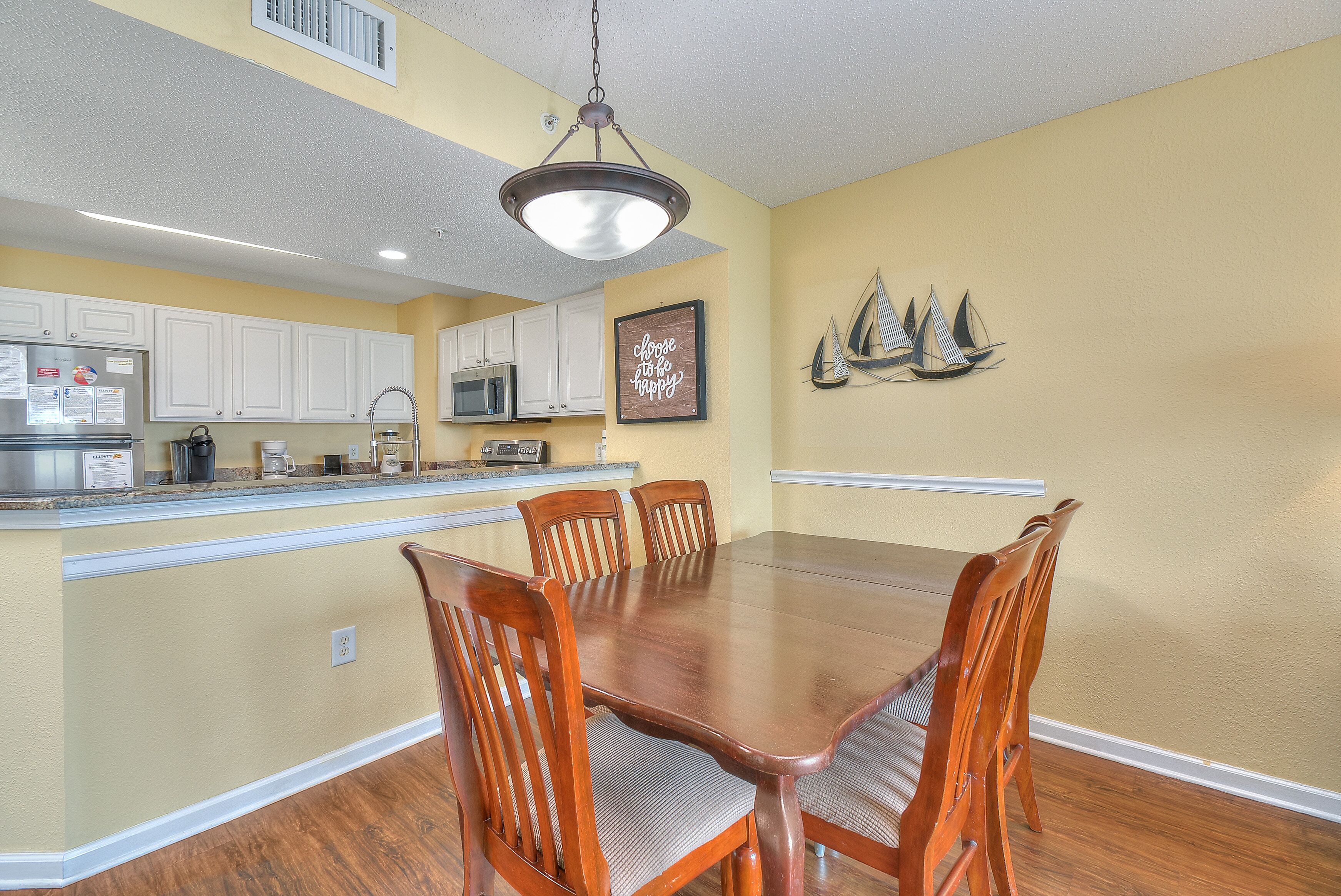 A dining area with a wooden table and chairs, a kitchen in the background, and a decorative sailboat on the wall.