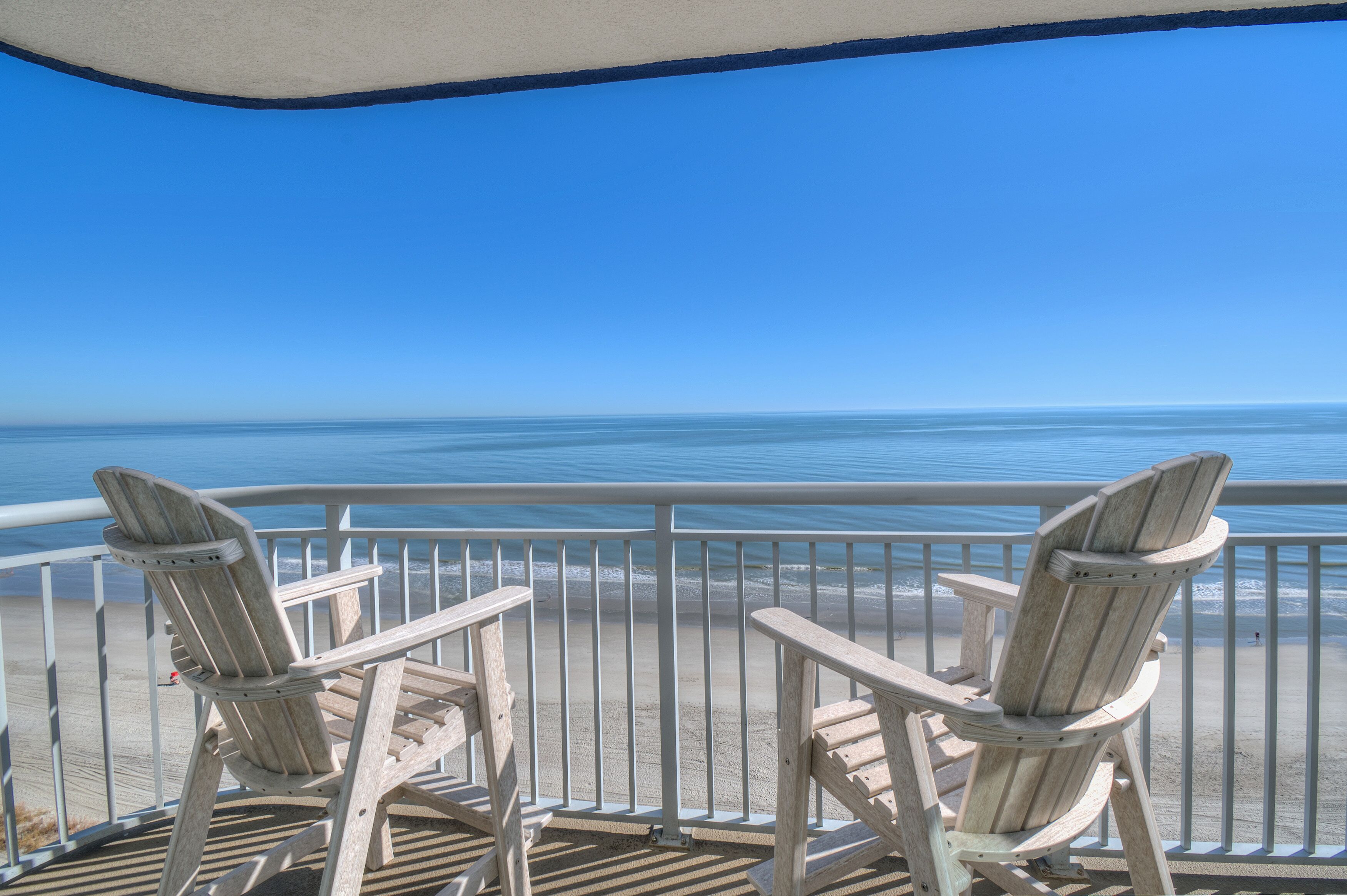 Two wooden Adirondack chairs on a balcony overlooking a beach with a clear blue sky.