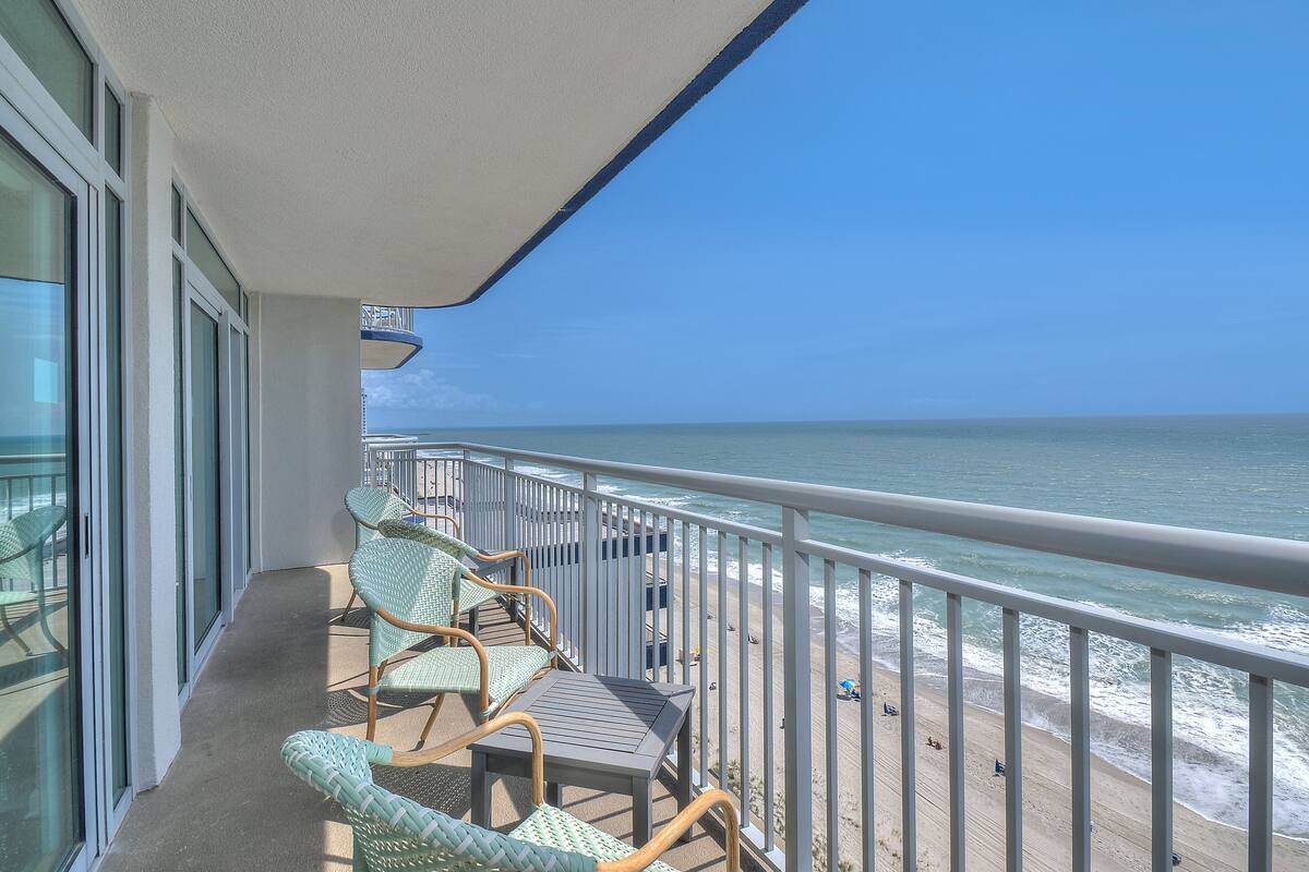 Ein Balkon mit Korbstühlen und einem Tisch mit Blick auf einen Strand, an dem sich Menschen befinden und ein Rettungsturm steht.