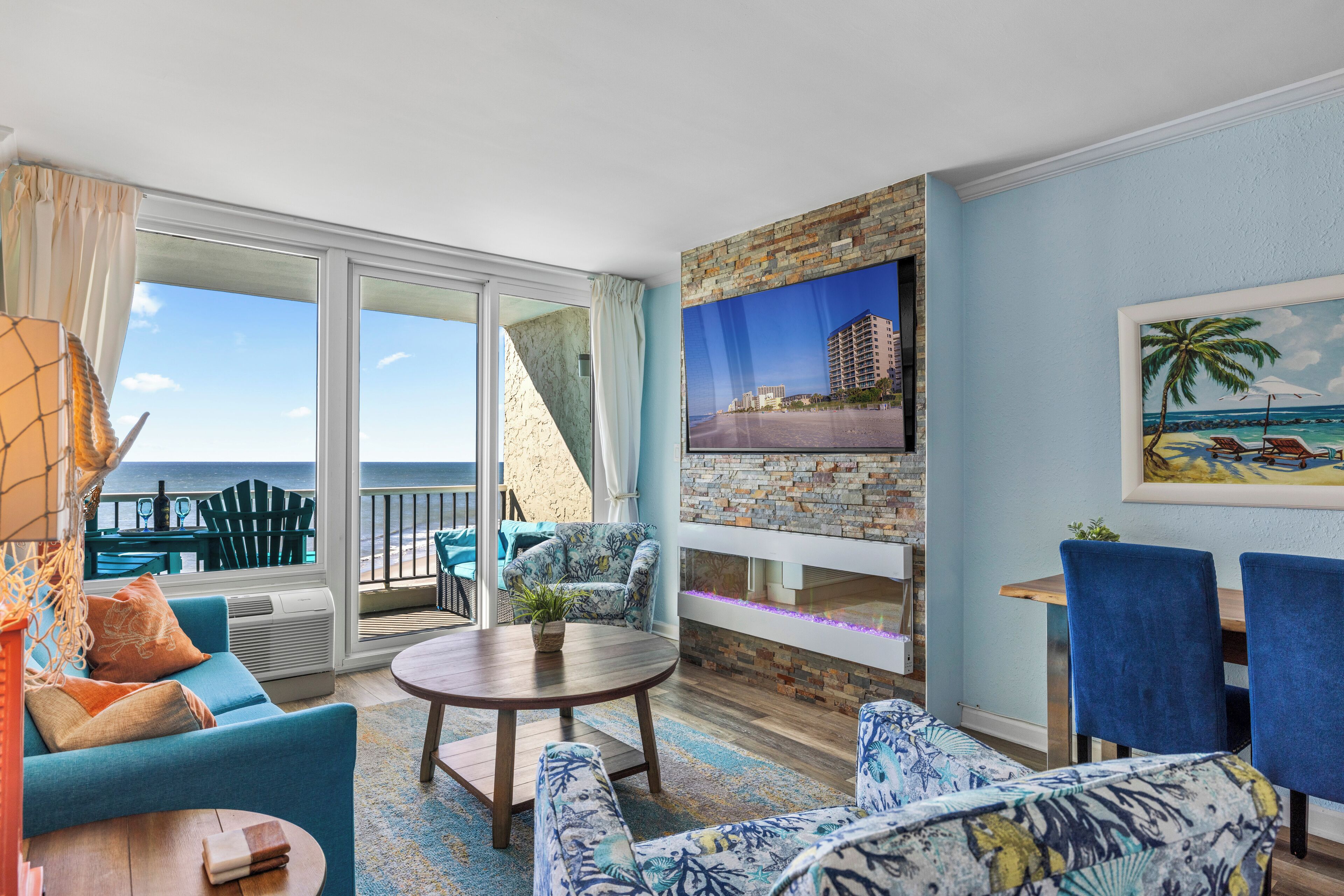 A spacious living room with a large window overlooking the ocean, a stone fireplace, and a view of a hotel building.